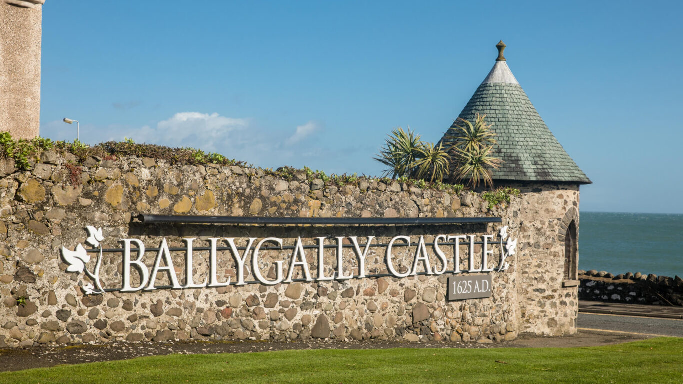 A stone wall featuring a turret has in large letters: " Ballygally Castle, 1625 AD" on a sunny day with the blue sea in the background.