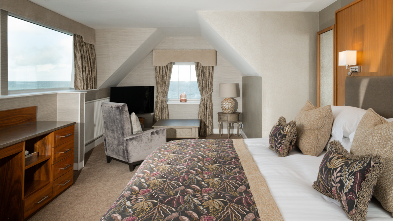 Elegant hotel room with sea view, plush bedding, cosy armchair, sleek desk, flat-screen TV, and warm lighting.