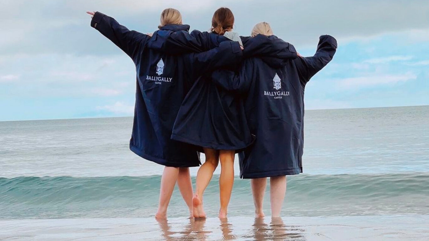 Three women wearing plush Ballygally Castle bathrobes stand in the ocean on a bright day