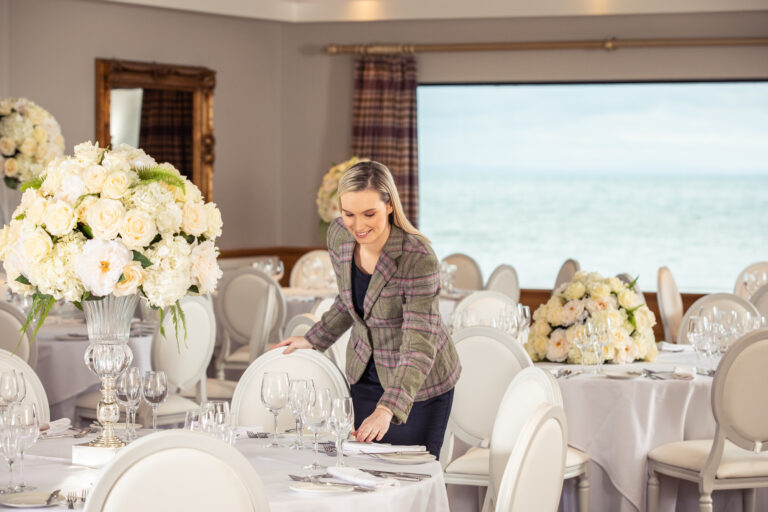A smartly dressed host fixes the table settings on a beautifully laid table covered in white linens, sparkling glasses and large bouquets of white flowers