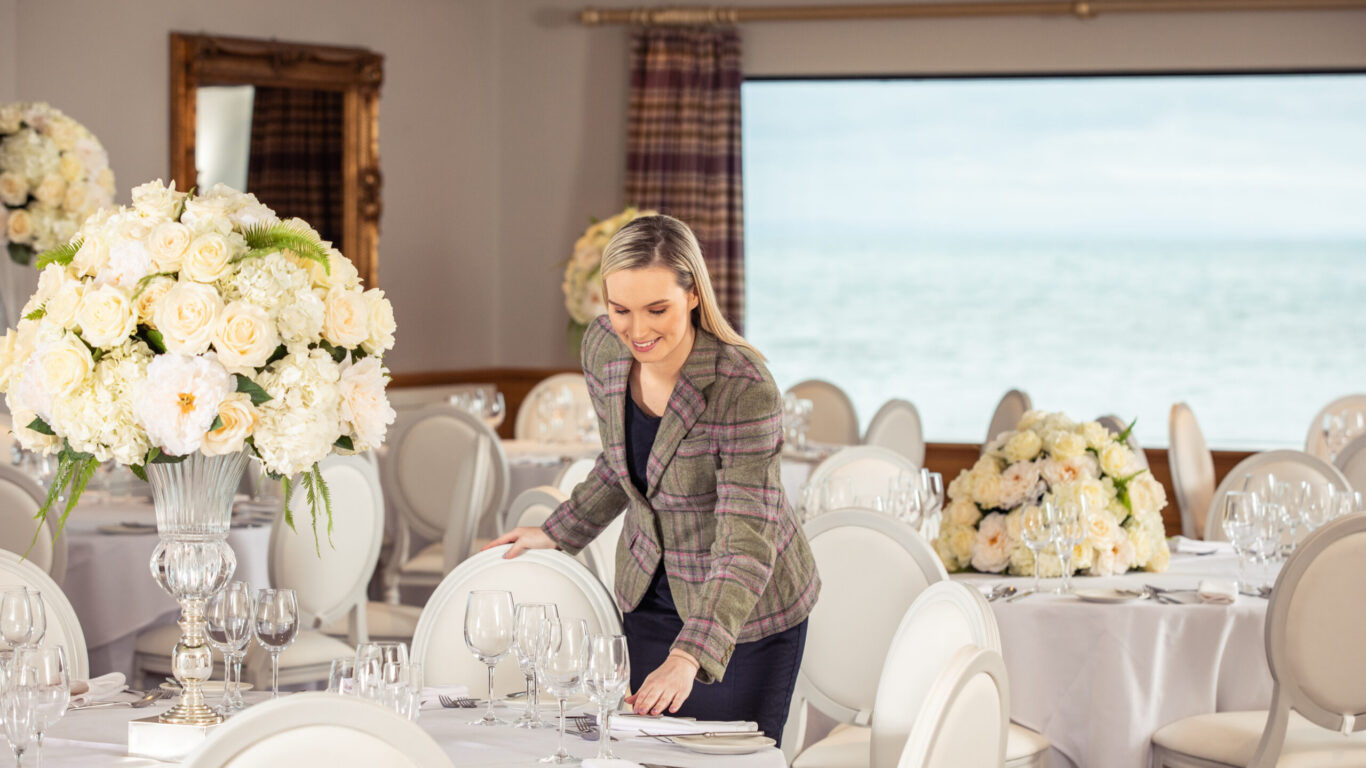 A smartly dressed host fixes the table settings on a beautifully laid table covered in white linens, sparkling glasses and large bouquets of white flowers