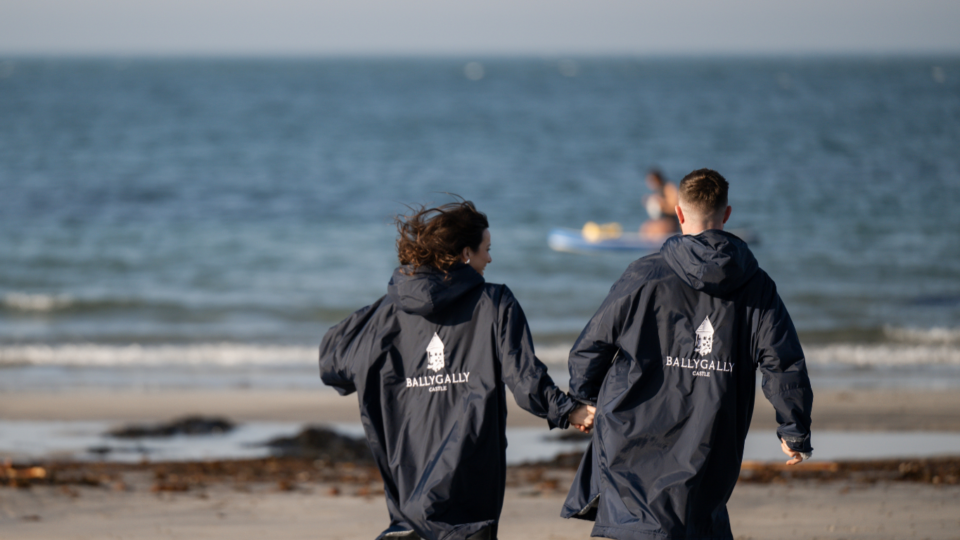 Couple holding hands on serene beach, wearing branded Ballygally Castle jackets, enjoying the coastal view.