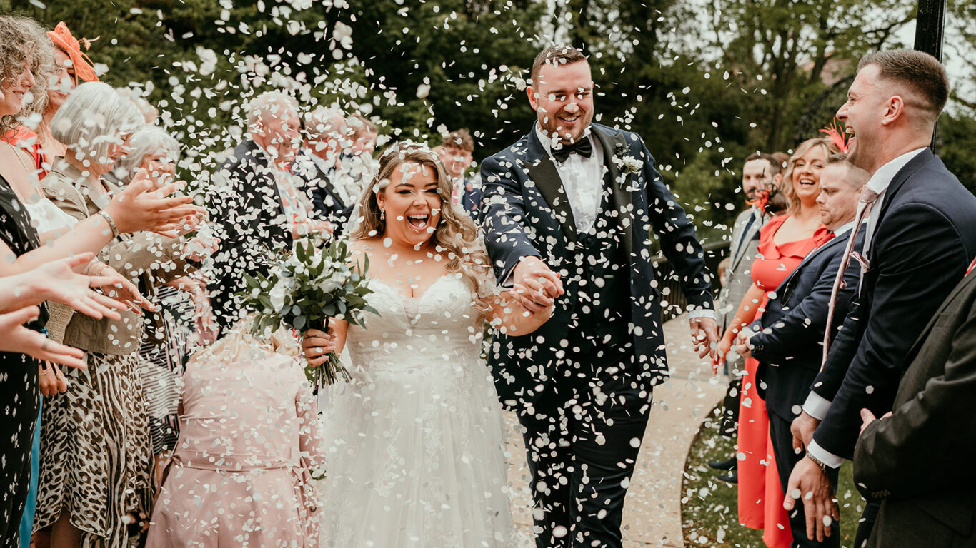 Joyful wedding couple walk through confetti amidst cheering guests in lush garden setting.