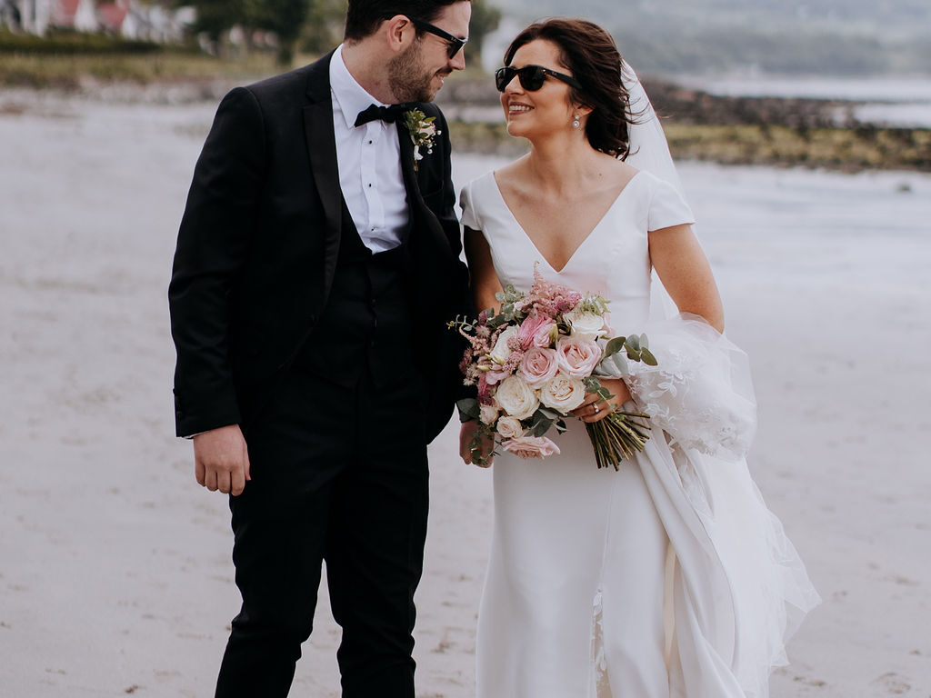 Bride and groom smiling while walking on a scenic beach, with rolling hills and charming cottages in the background.