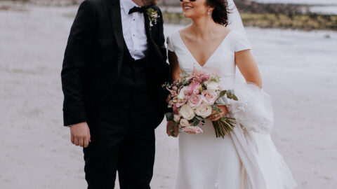 Bride and groom smiling while walking on a scenic beach, with rolling hills and charming cottages in the background.