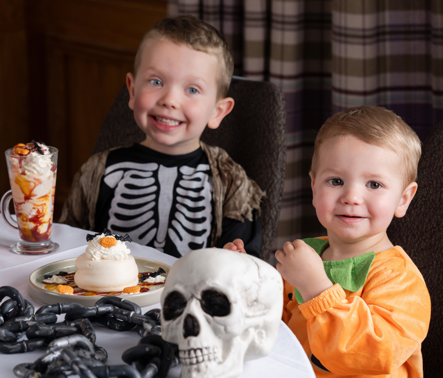 Children in Halloween costumes enjoy desserts at a festively decorated hotel restaurant table, creating a joyful atmosphere.