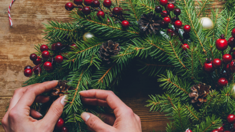 Hands crafting a festive wreath with pinecones and berries on a wooden table at a cosy hotel, perfect for a holiday retreat.