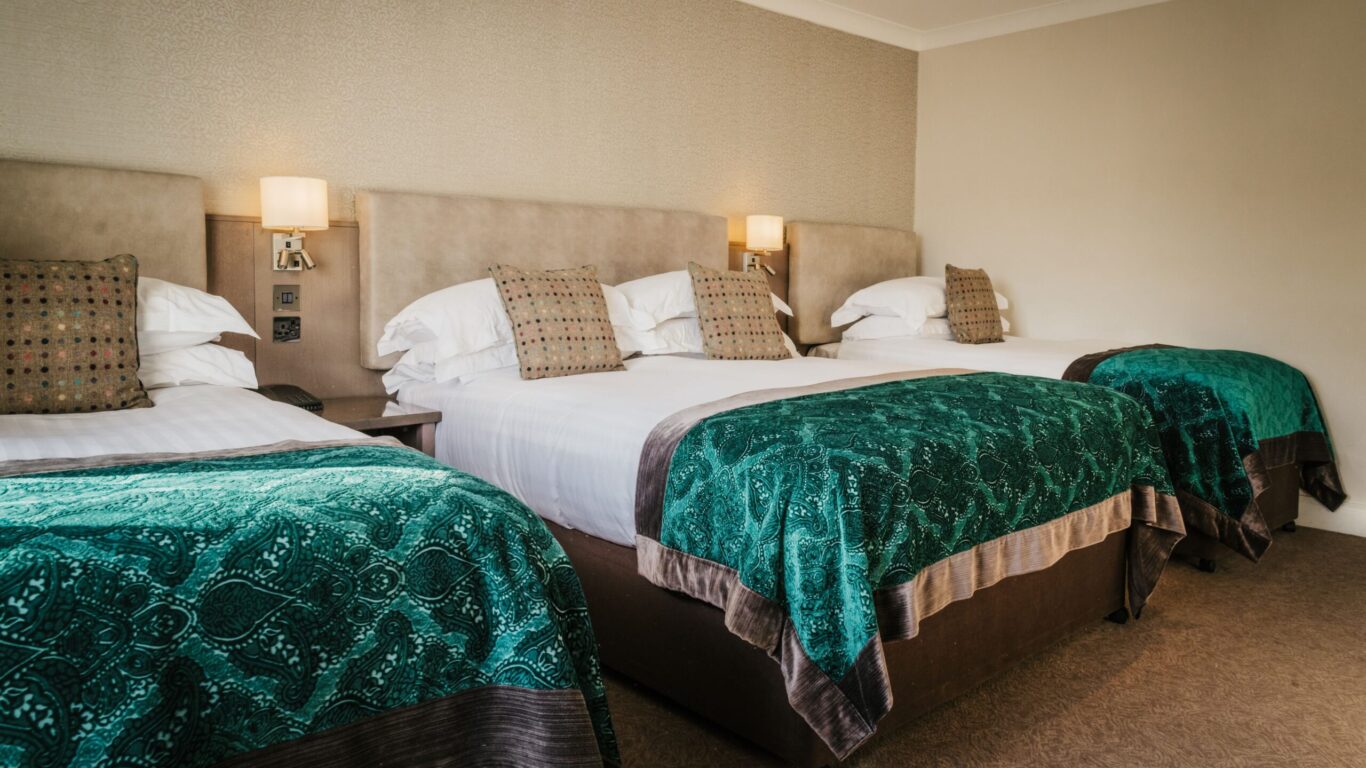 Three neatly made hotel beds with teal-patterned blankets and multiple pillows, positioned side by side in a neutral-toned room with wall-mounted lamps.
