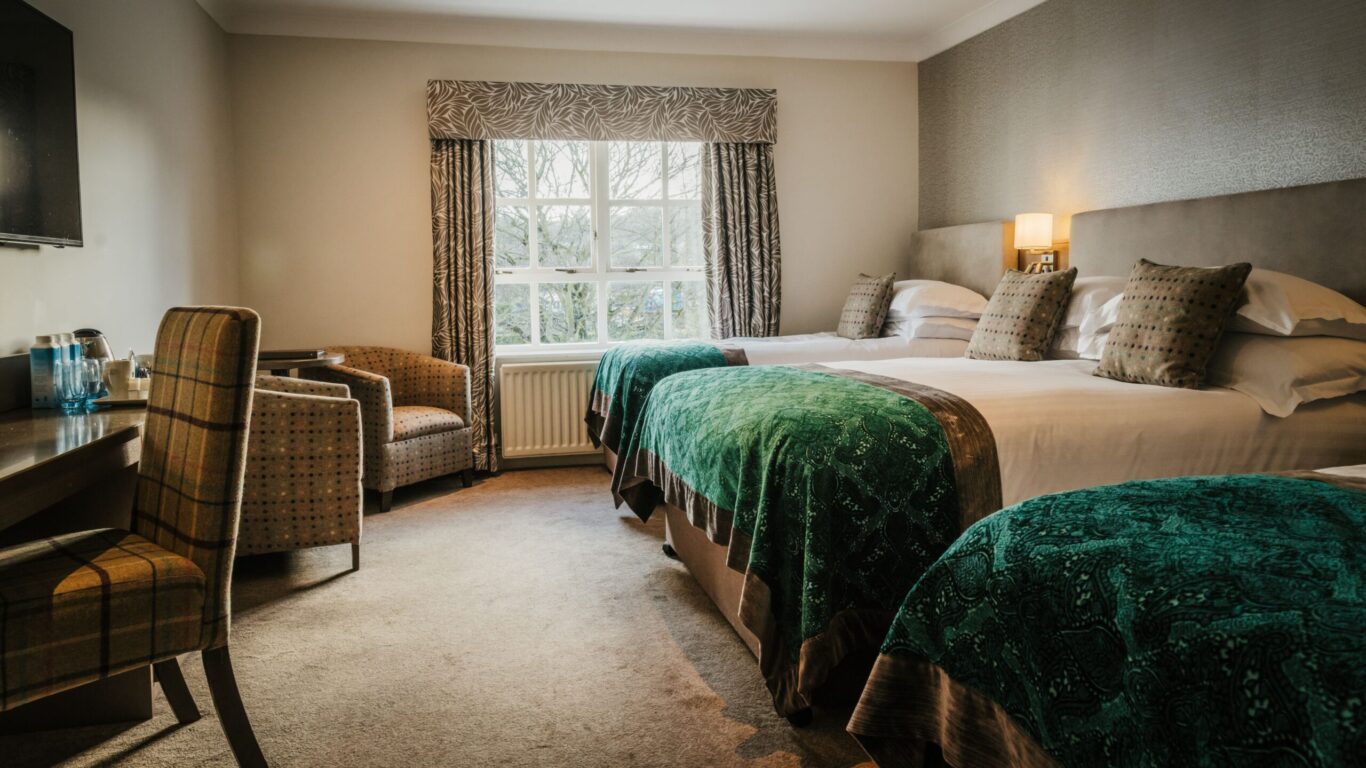 A hotel room with two double beds covered in green blankets, a desk with a chair, an armchair by the window, and neutral-toned walls and decor.