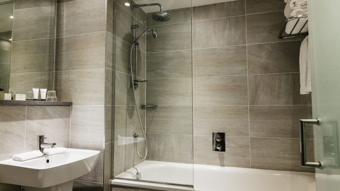 Modern bathroom with beige tiled walls, a glass-enclosed bathtub with a shower, a white sink, toiletries, and neatly stacked towels on a metal rack.