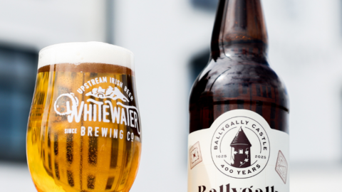 A glass of pale lager with foam sits beside a 500ml bottle of Ballygally Castle Crisp Lager, 4.3% ABV, on an outdoor surface.