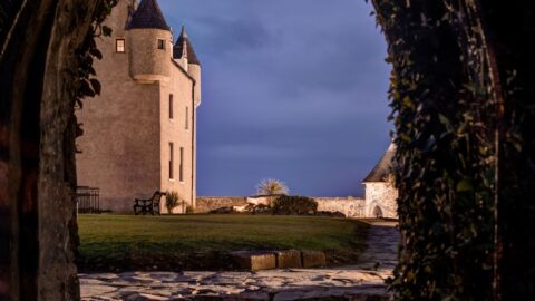 View through an arched stone doorway, looking out onto a lawn and a stone castle with turrets under a cloudy evening sky.