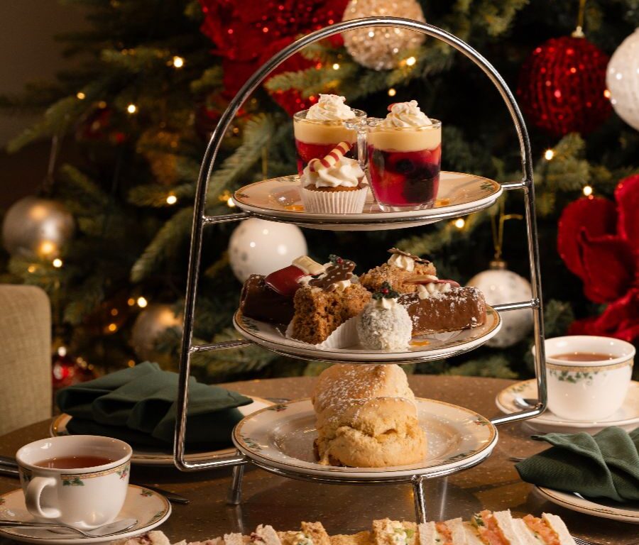 A three-tiered stand with assorted desserts and pastries, a plate of finger sandwiches, two cups of tea, and a decorated Christmas tree in the background.