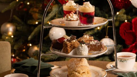 A three-tiered stand with assorted desserts and pastries, a plate of finger sandwiches, two cups of tea, and a decorated Christmas tree in the background.