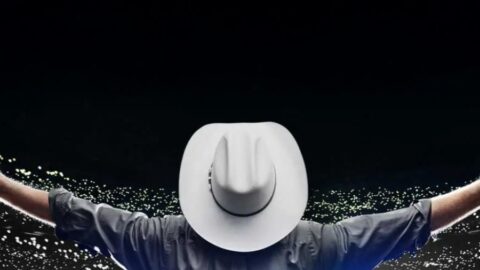 A person in a cowboy hat stands on stage with arms outstretched, facing a large audience with illuminated lights in the background.