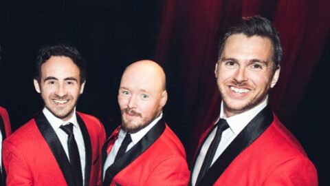 Three men wearing matching red blazers with black lapels, white shirts, and black ties stand together and smile at the camera against a dark background.