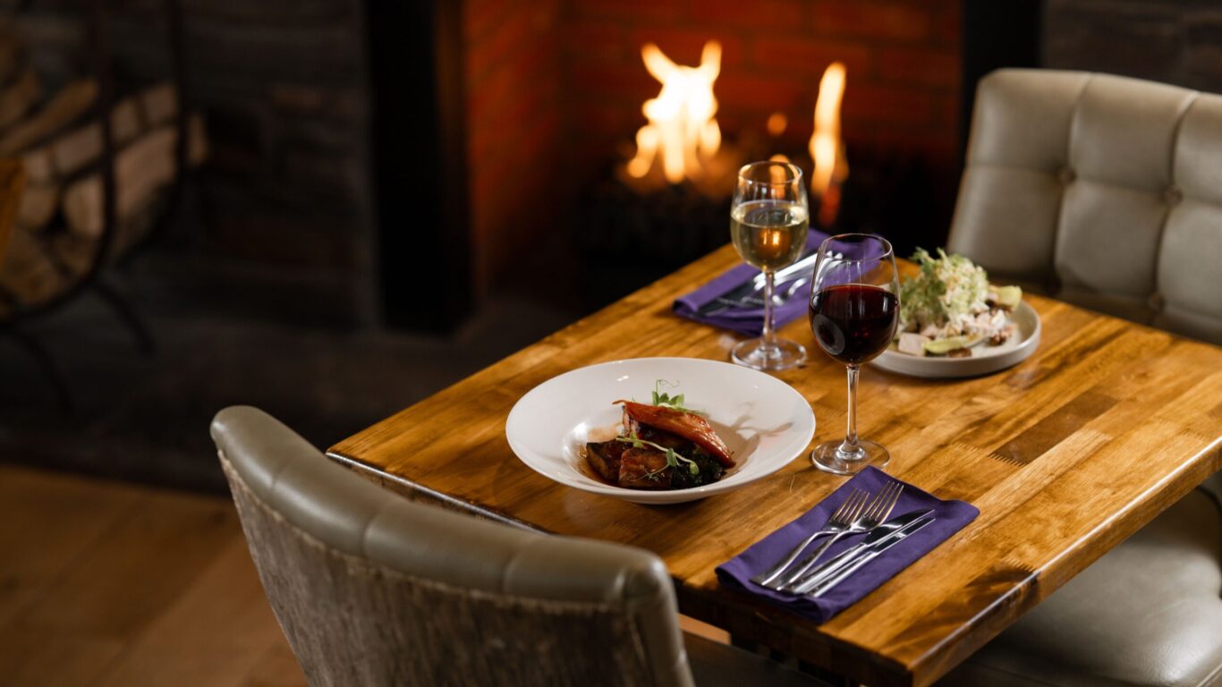 A wooden table set for two with plated meals, glasses of red and white wine, and cutlery, positioned by a lit fireplace.