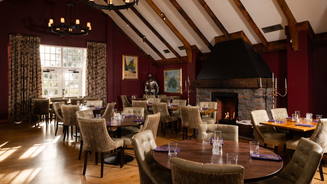 Upscale restaurant dining room with tufted chairs, round tables set with glassware, a large stone fireplace, sloped wooden beam ceiling, and natural light from a window.