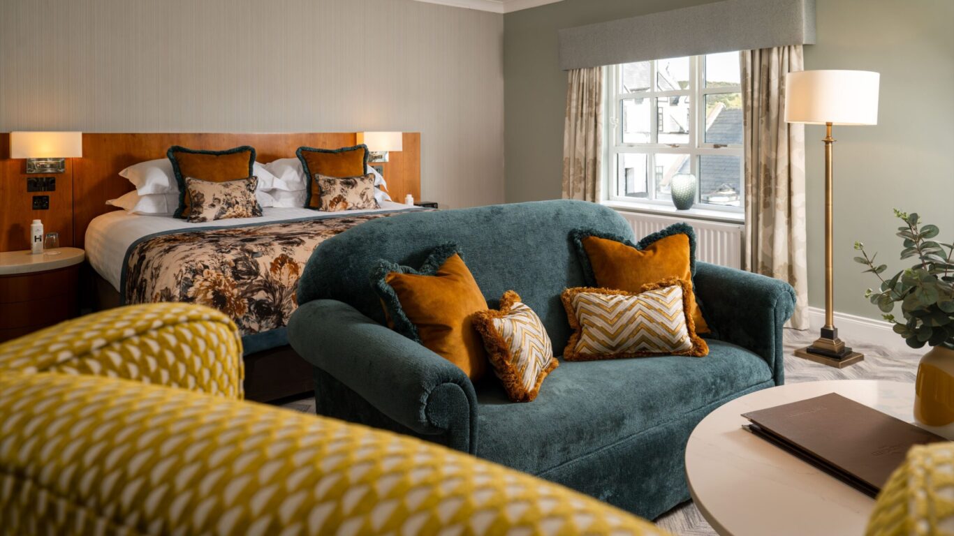 Hotel room with a double bed, green sofa, yellow and patterned cushions, a round table with folders, and a window with curtains letting in natural light.