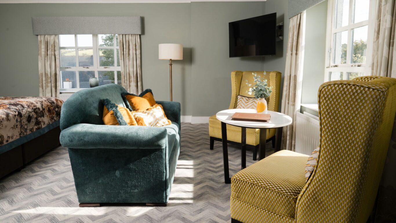 A living area with a green sofa, two yellow armchairs, a round white table, a bed, floor lamp, wall-mounted TV, and large windows with curtains.